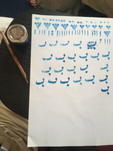 A jar of ink, reed pen, and a sheet of paper with Arabic calligraphy letters