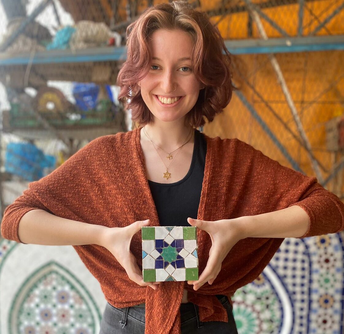 Female student holding colored mosaic tile with a wall of mosaic tiles behind her in Morocco.