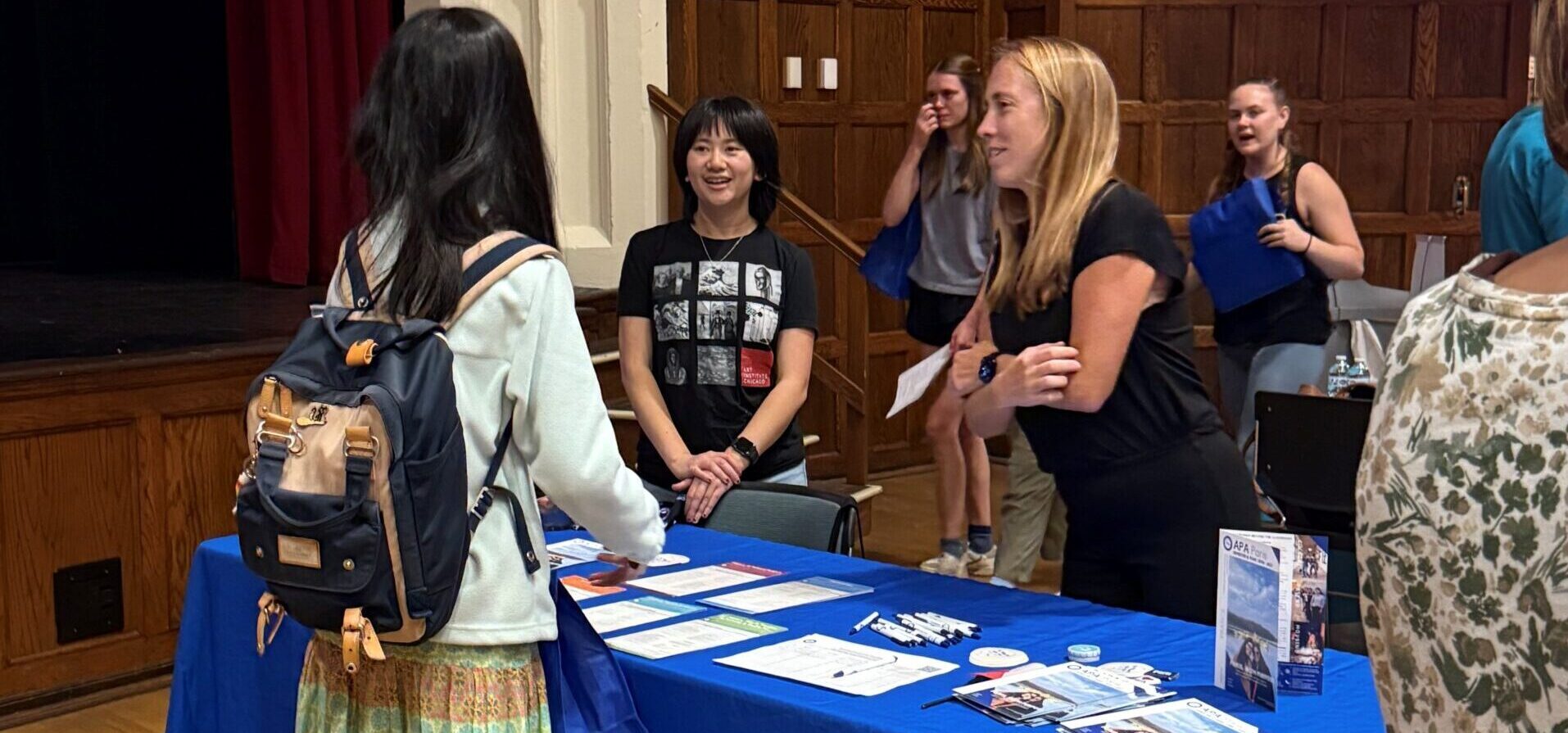 2 females, APA alum and Staff member, behind a table speaking with students at a study abroad fair.